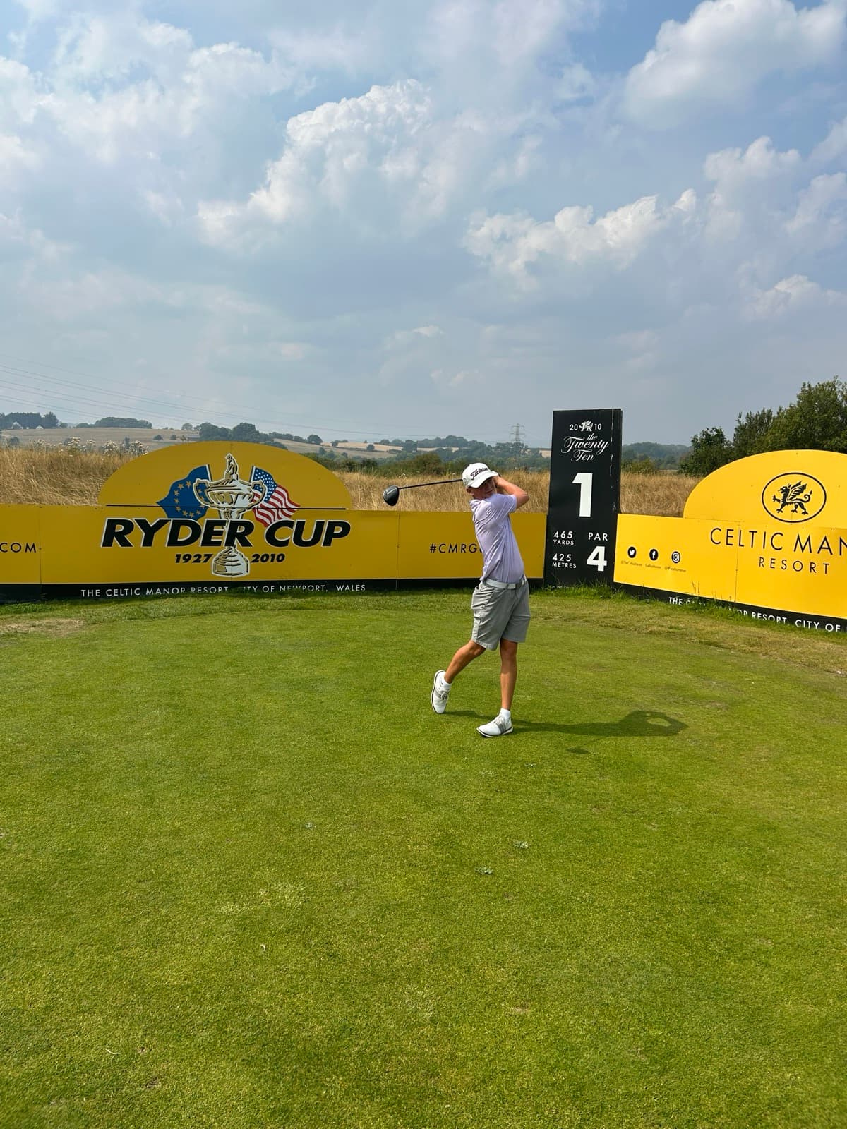 Sam Eynon at the top of his backswing with a driver on the first tee of the Twenty Ten course at Celtic Manor, Ryder Cup signage behind him
