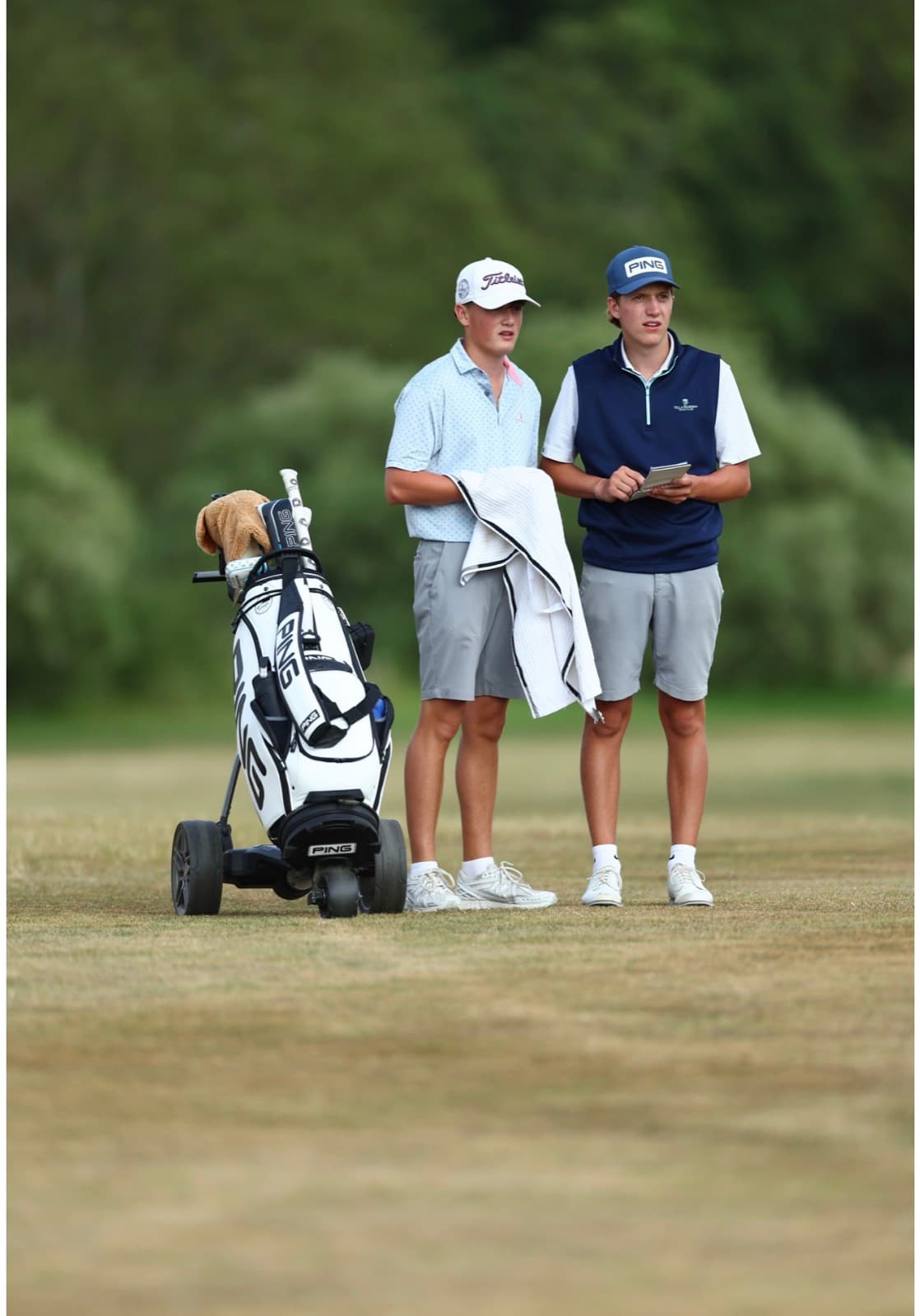 Sam Eynon standing on a fairway with a towel over his arm beside a Ping bag on a trolley, another player checking a yardage book next to him