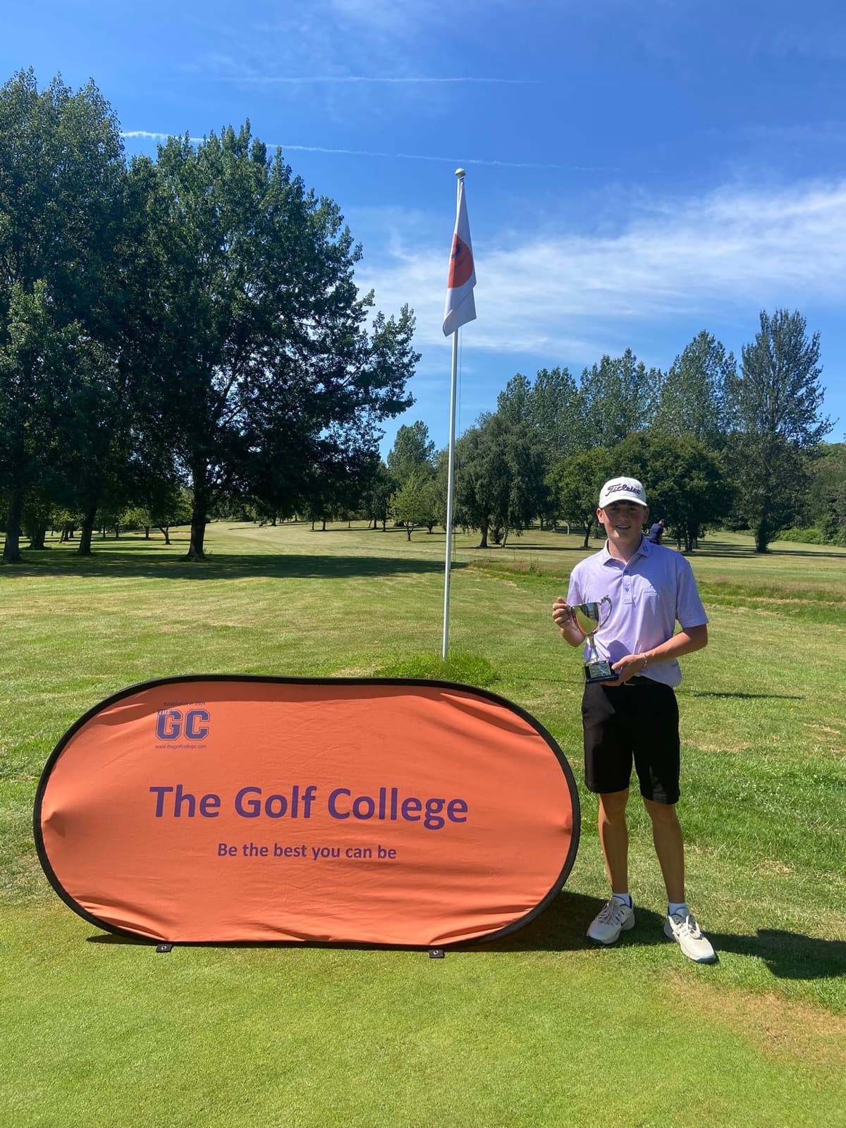 Sam Eynon holding a silver cup beside a The Golf College banner on a golf course