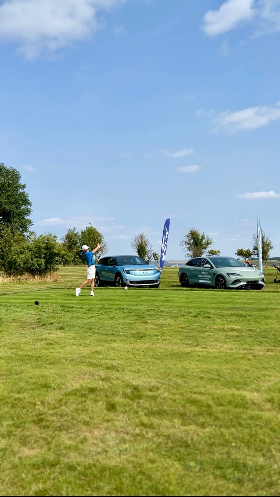 Sam Eynon following through a tee shot on a downland course, two display cars and banner flags beside the tee
